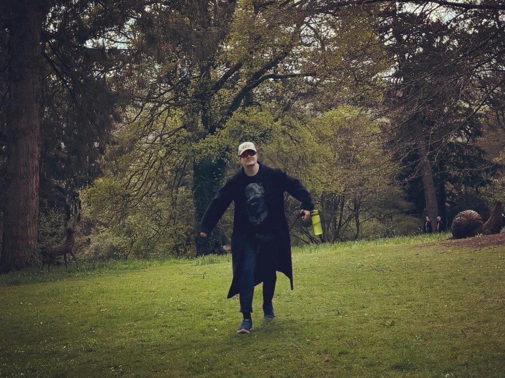 Harrison walking towards the camera wearing a long black coat and with his arms wide.