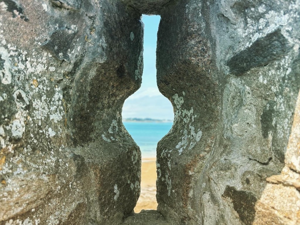 Vertical slit in a stone wall with the sea and beach beyond.